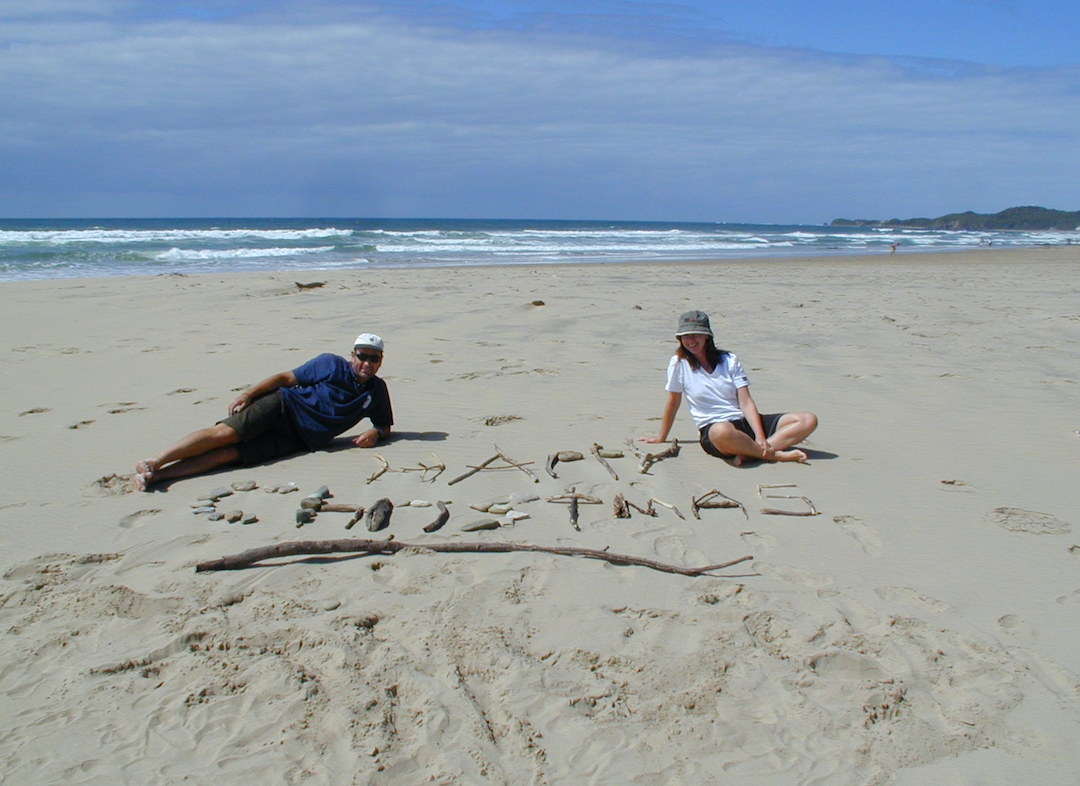 David and Sharon Schindler lying on a beach with Happy Christmas written in the sand
